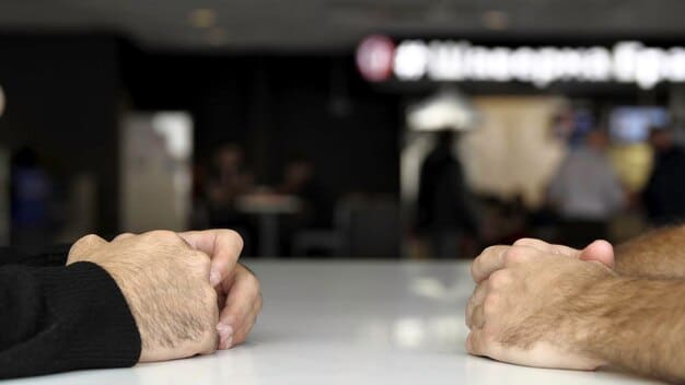 A close-up shot of a clenched fist resting on a table, with a slightly blurred background of two people arguing intensely. The lighting is dim and reflects the stress of the situation.