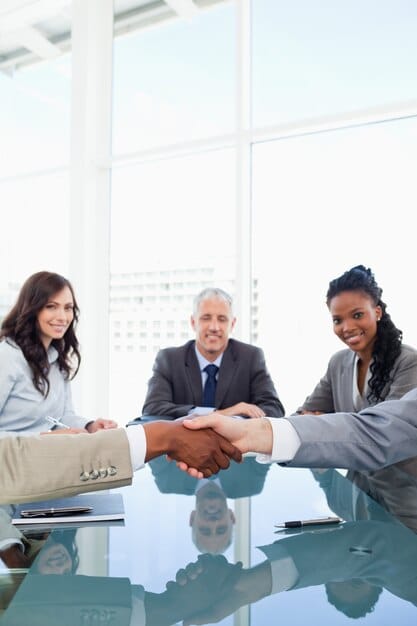 Two business leaders shaking hands across a boardroom table, with a diverse team visible in the background, symbolizing successful strategic partnership and collaboration.